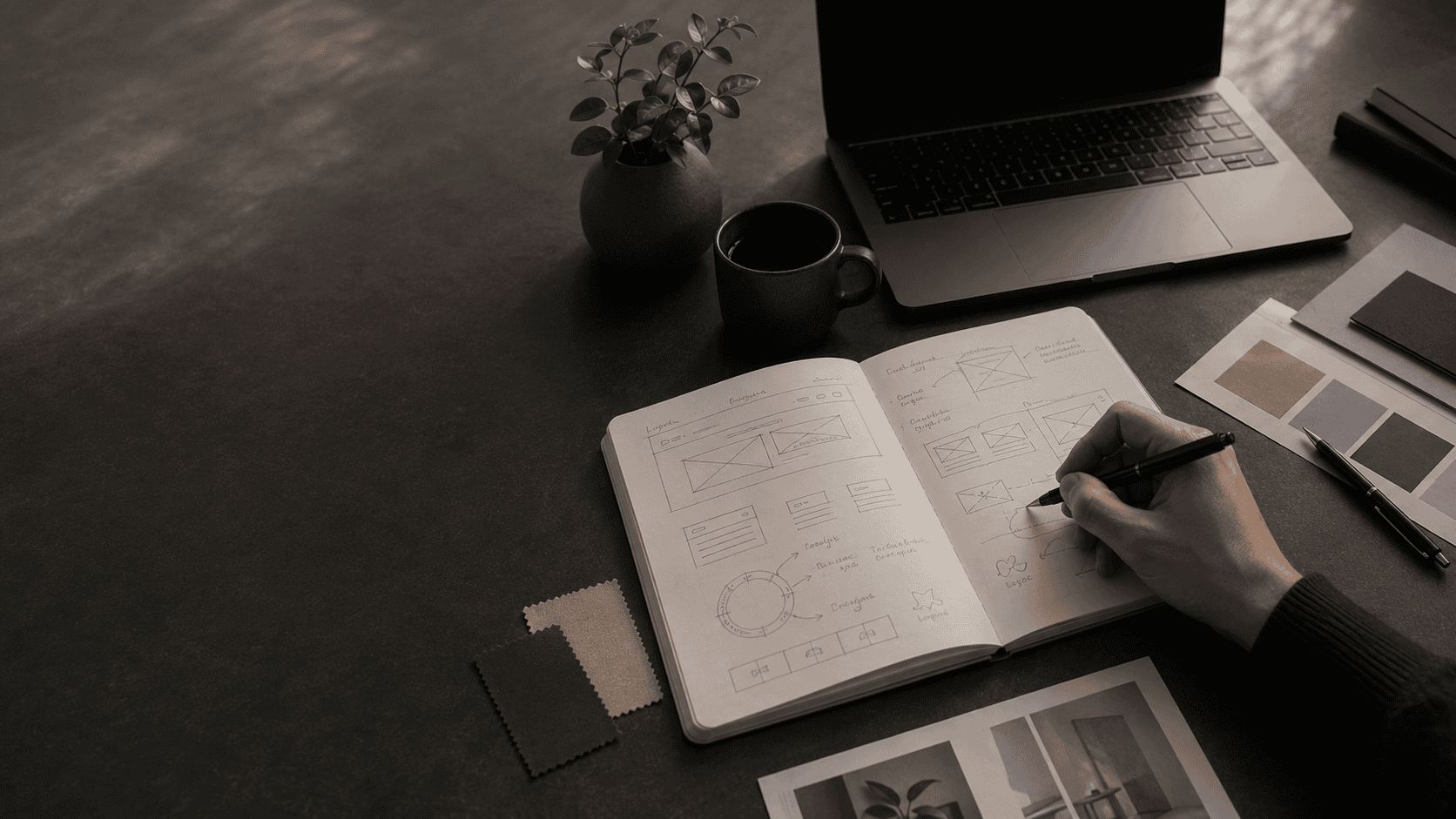 Minimal brand strategy workspace with a laptop, open notebook, color palette samples, printed design materials, coffee cup, and plant on a dark desk in natural light.