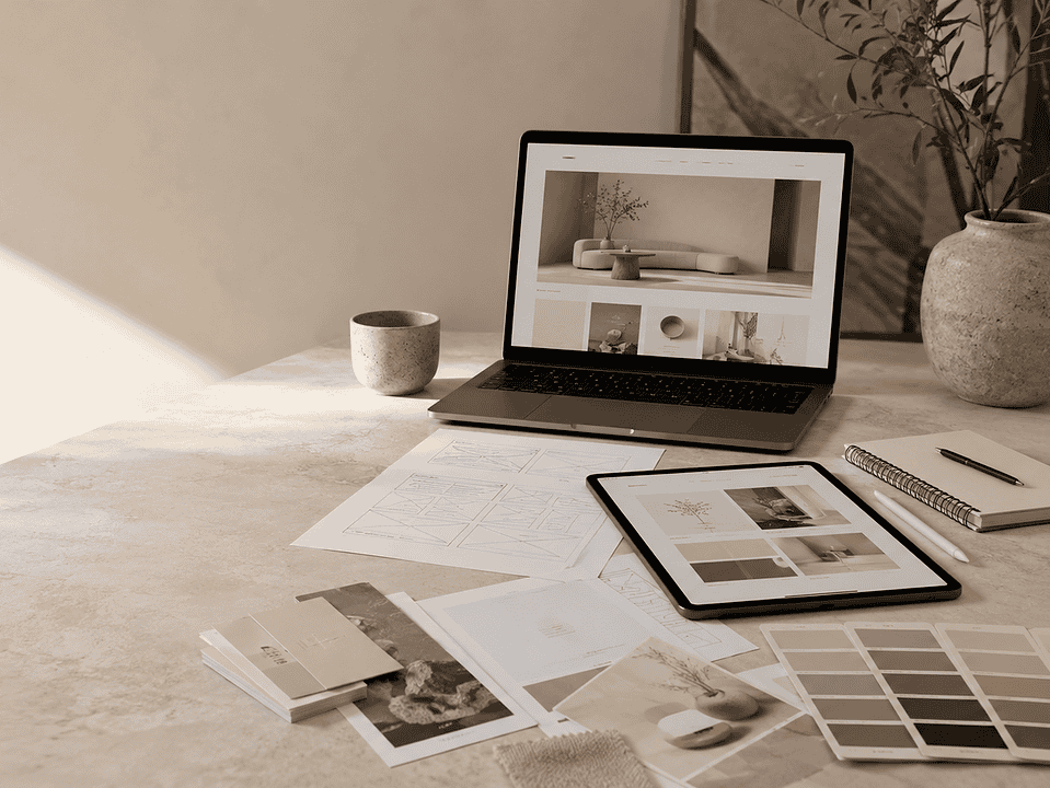 A workspace with a laptop and tablet displaying website designs, surrounded by wireframe sketches, color swatches, notebooks, and branding materials in a soft, natural light setting.